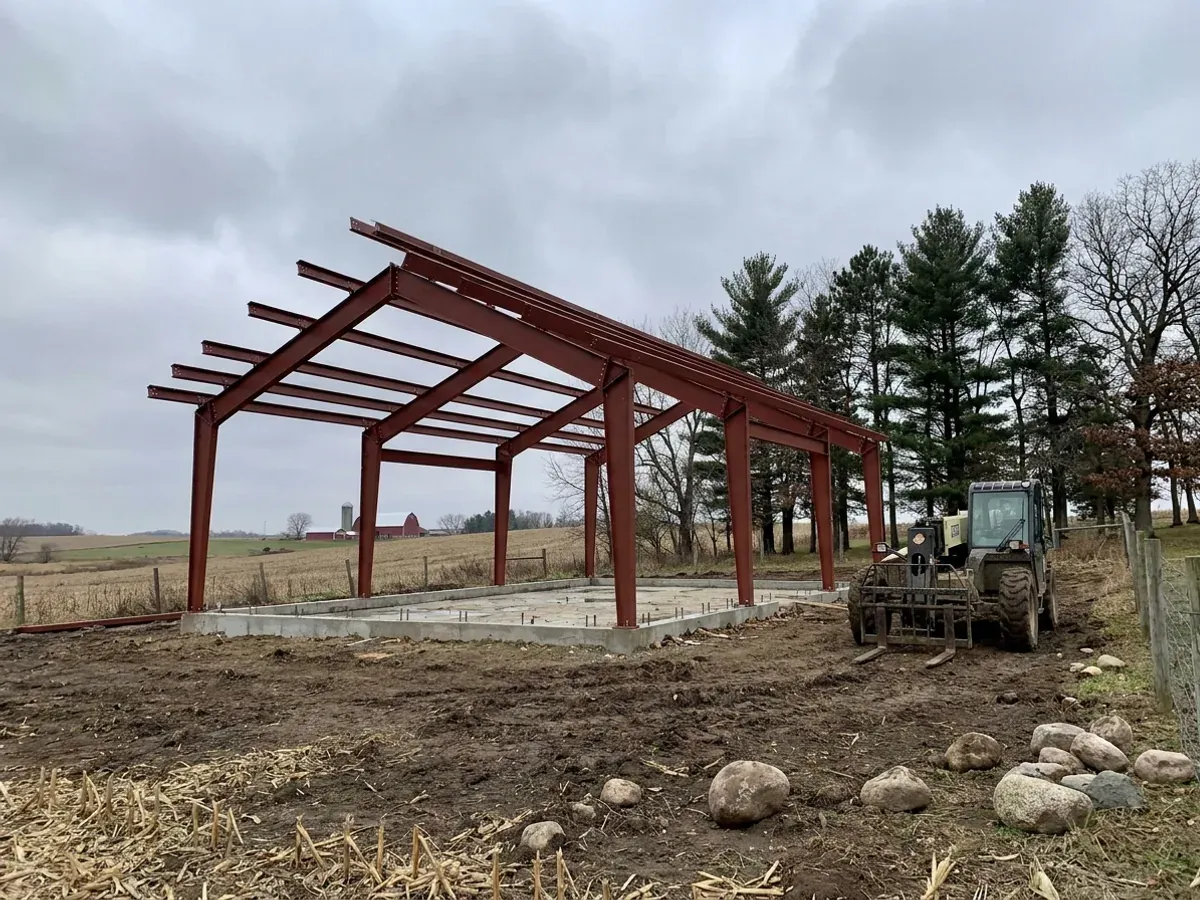 A red oxide primed structural steel frame partly erected on a concrete slab, showing bolted knee joints and anchor bolts.