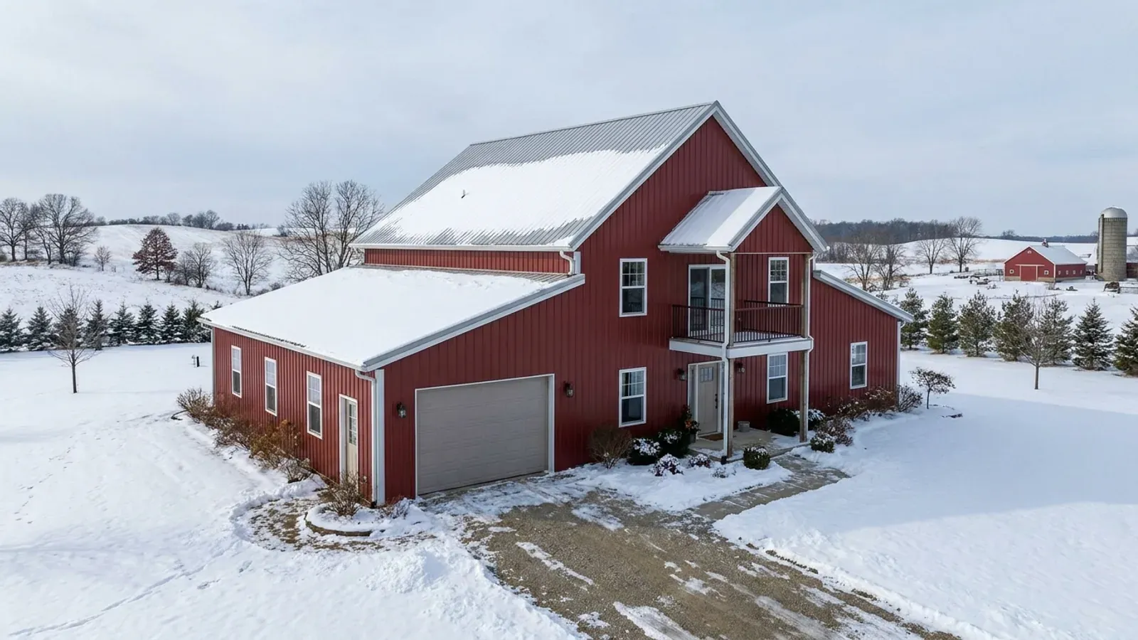 A barn-red two-story barndominium in winter with snow on its steep roof, second-story windows and a small balcony above the entry.